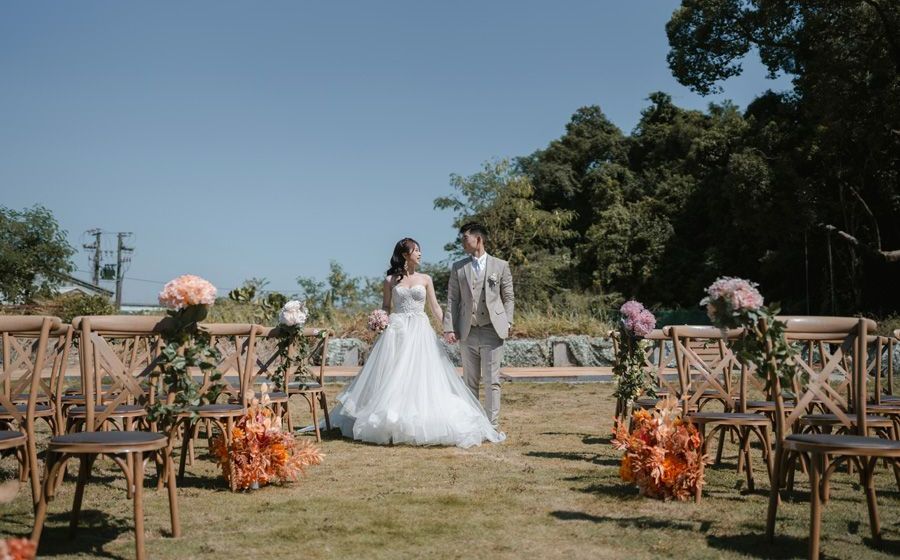 Bride and groom stand in outdoor wedding aisle, holding hands. Chairs are decorated with flowers.