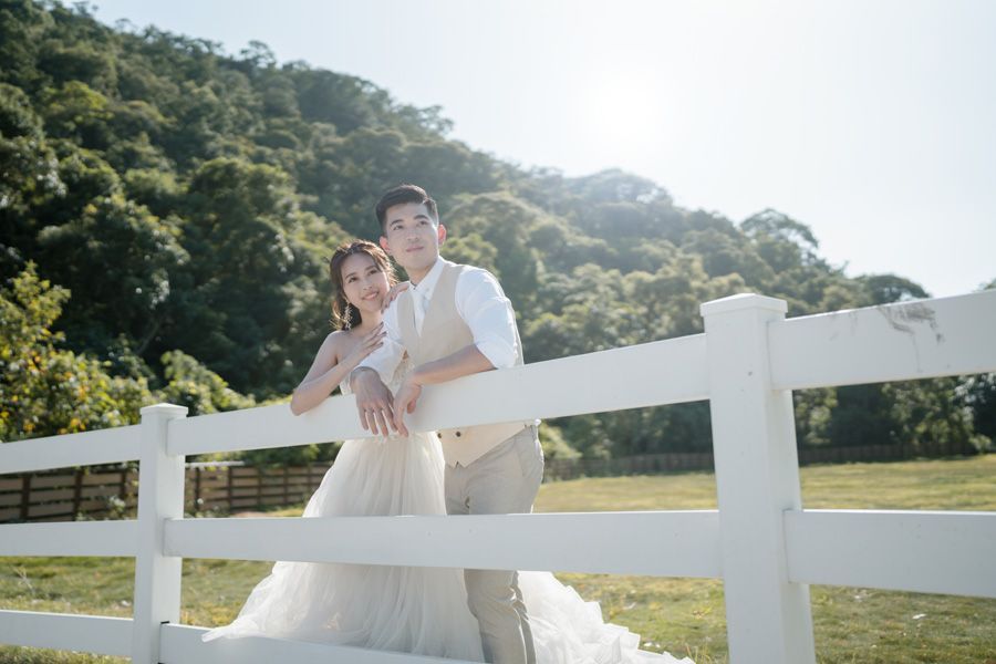 Couple in formal attire leans against a white fence, posing in front of a green hillside.