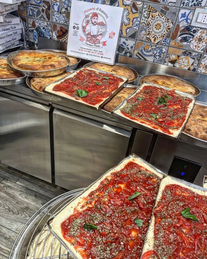 Vassoi di pizza su un bancone in una pizzeria, con una parete piastrellata a motivi decorativi sullo sfondo.