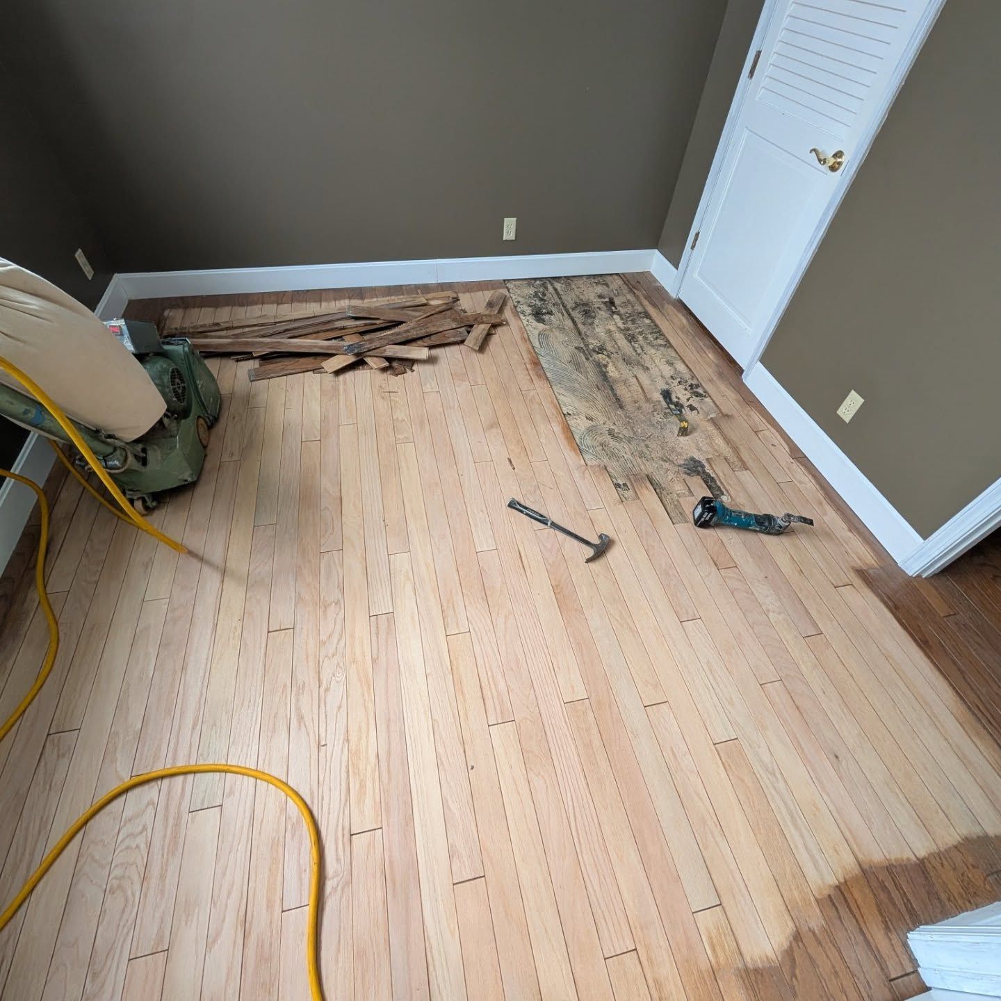 Partially repaired hardwood floor in a room, with tools and exposed underlayment near a wall.