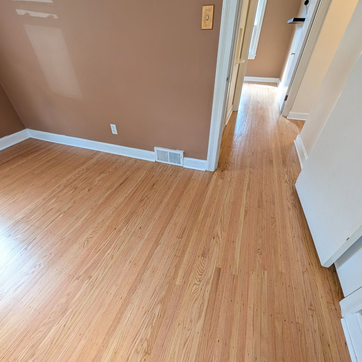 Empty room with light wood floors, brown walls, white trim, and a hallway doorway.