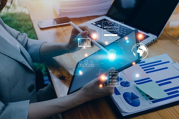 Woman using tablet with digital icons, laptop, charts, and papers on a wooden desk.
