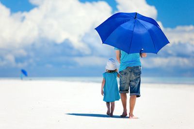Adult and child walk on a white beach under a blue umbrella on a sunny day.