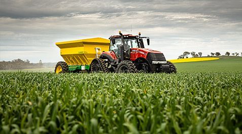 Red tractor with yellow fertilizer spreader in a field of green crops under a cloudy sky.