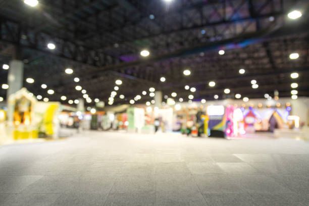 Blurred view of a large convention hall with booths, bright lights, and a gray floor.