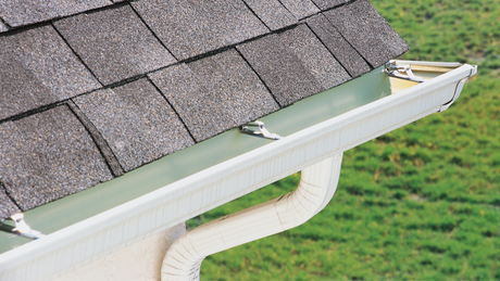 White gutter along the edge of a roof with gray shingles; a curved downspout leads away.