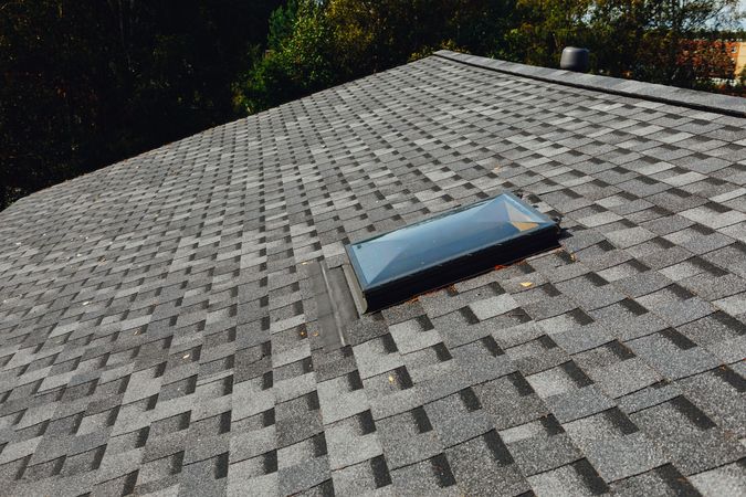 A rectangular skylight set into a gray, multi-tonal asphalt shingle roof on a bright, sunny day.