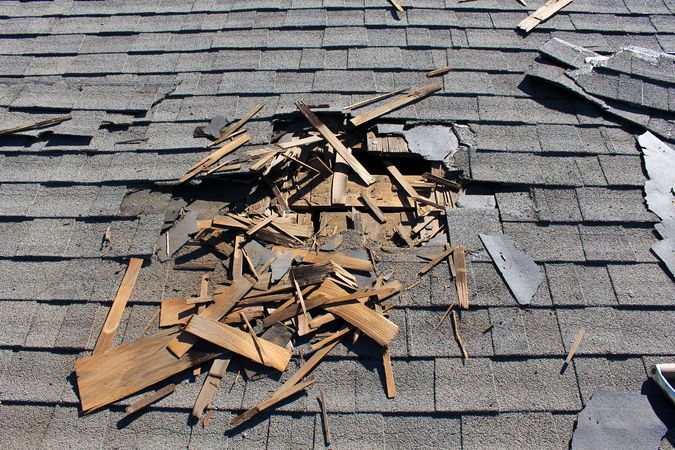 A damaged gray shingle roof with a hole exposing shattered wood decking and debris.