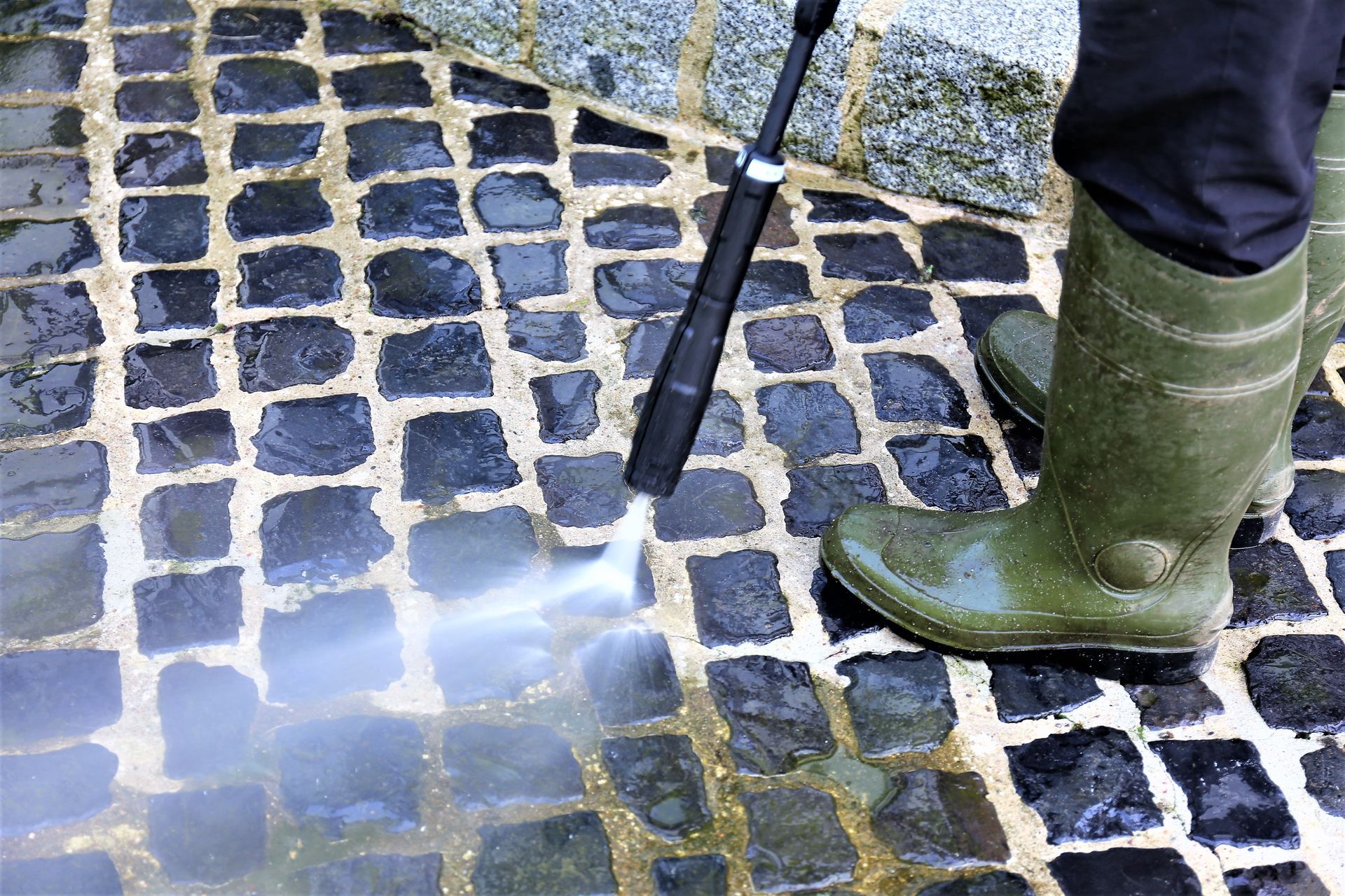 Green boots and pressure washer cleaning cobblestone pavement with water spray