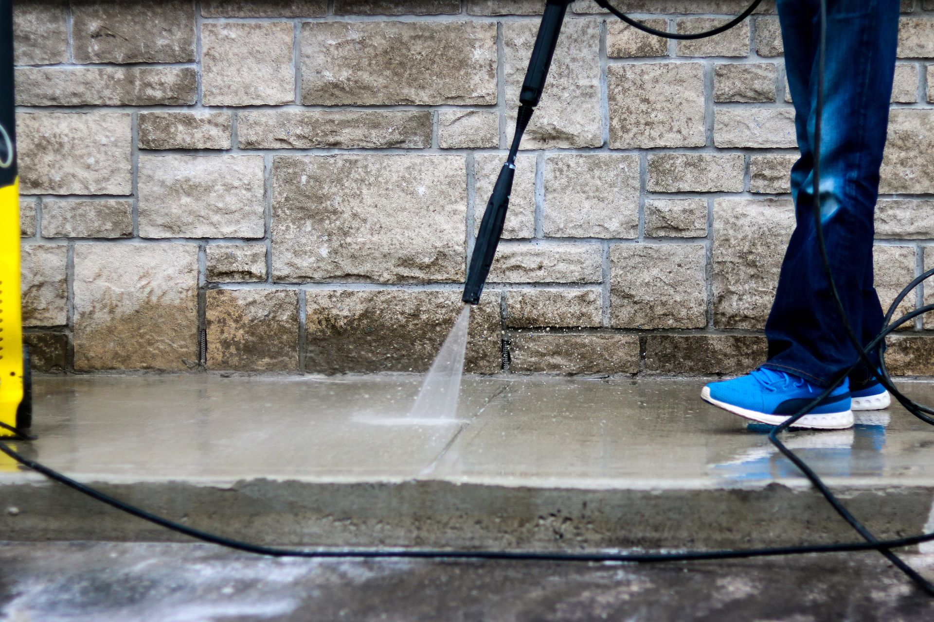 Person pressure-washing a stone wall and pavement; water sprays onto the wet ground.