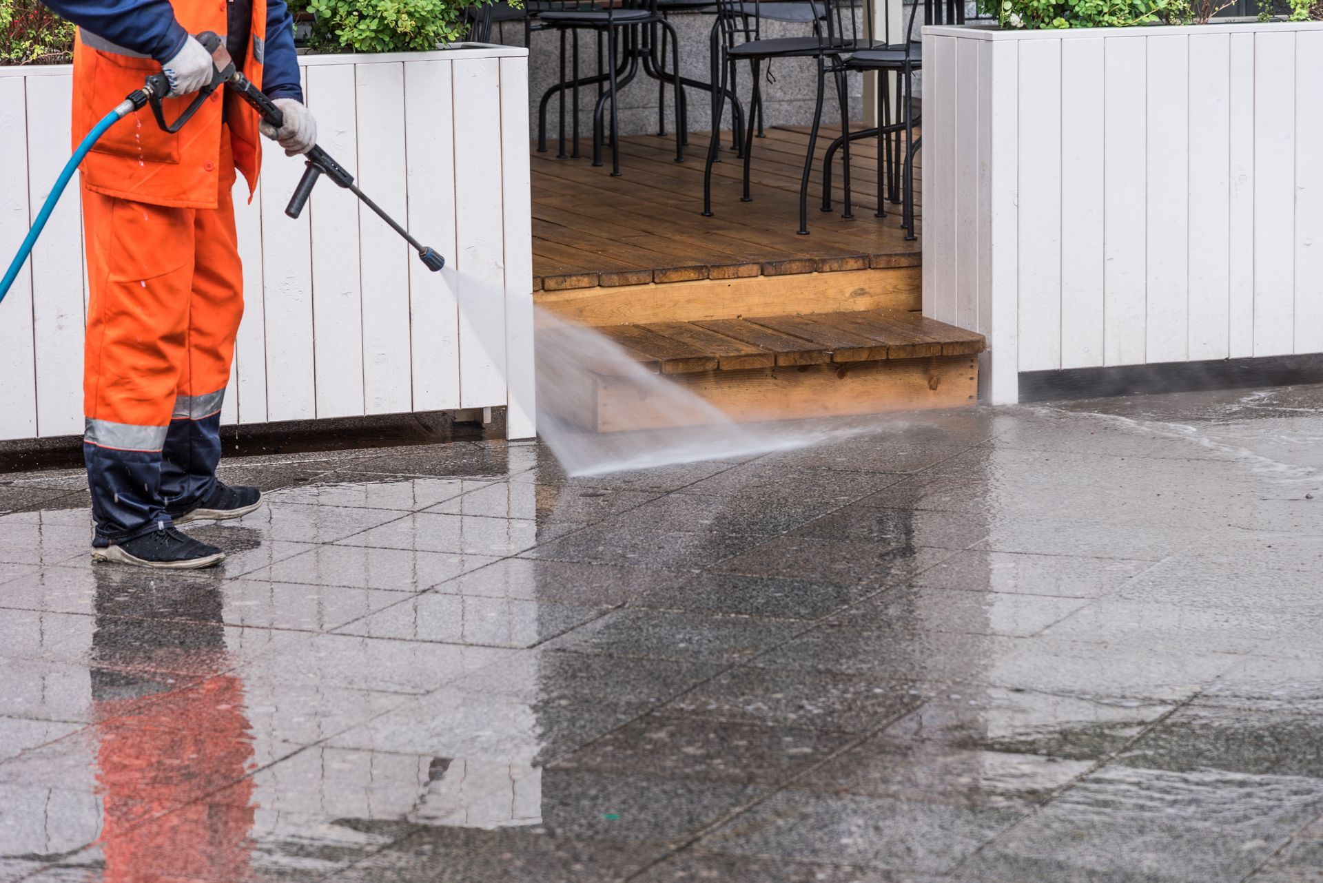 Worker in orange coveralls pressure-washing a wet sidewalk beside a white fence and outdoor café seating