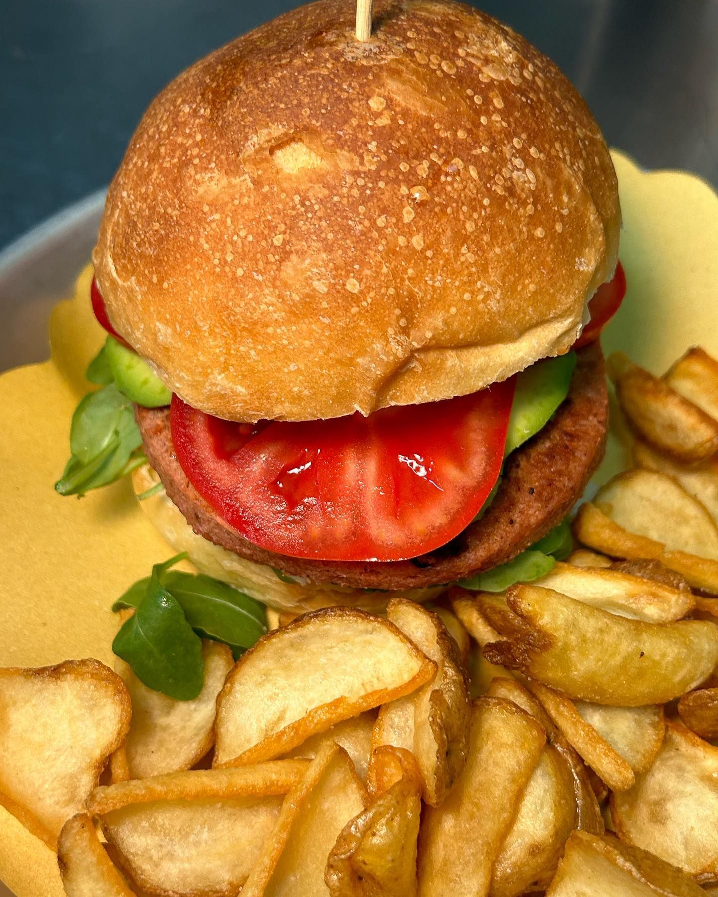 Hamburger in un panino con pomodoro, verdure e avocado, servito con un contorno di spicchi di patate.