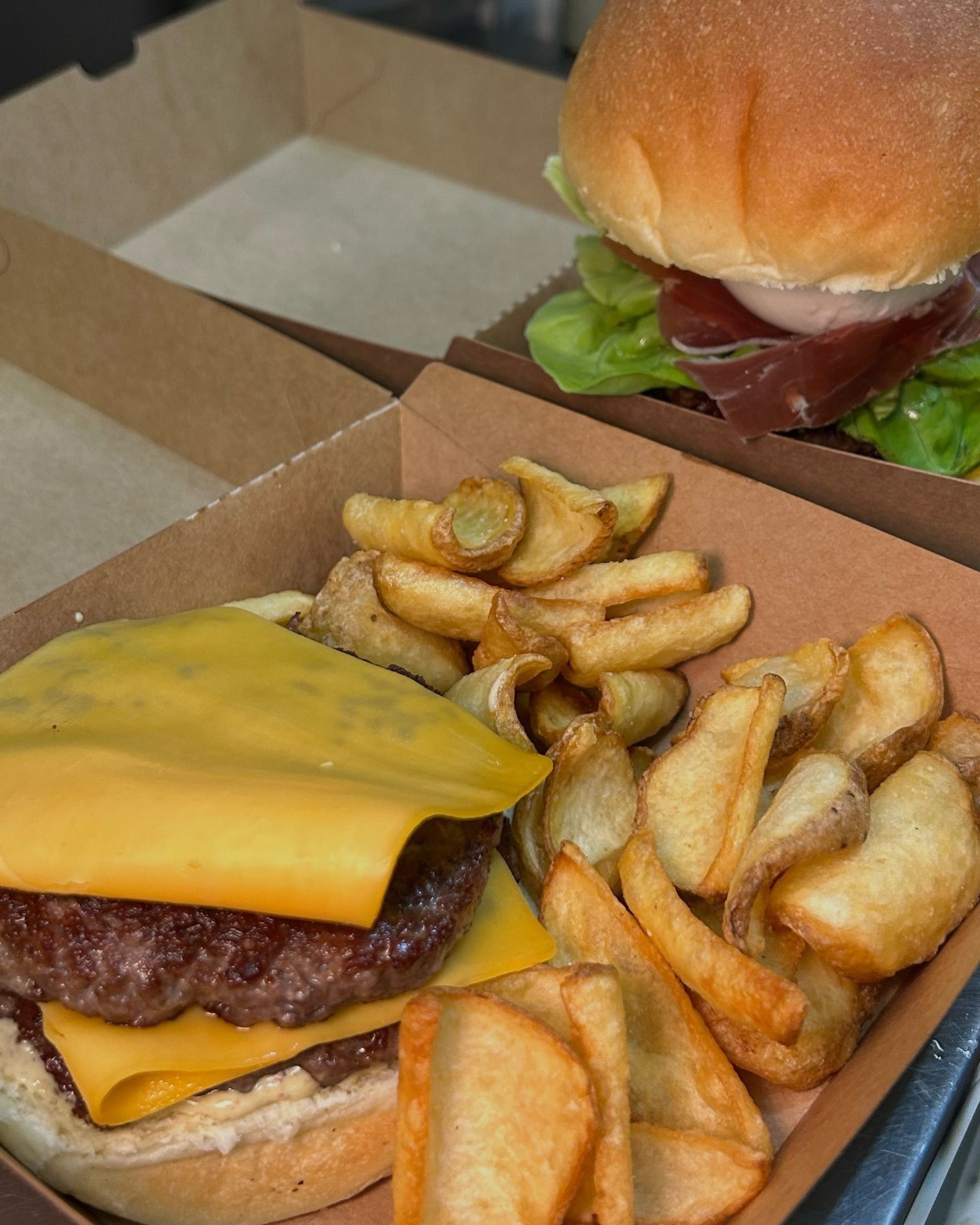 Cheeseburger e hamburger con lattuga e prosciutto in contenitori di cartone marrone con spicchi di patate fritte.