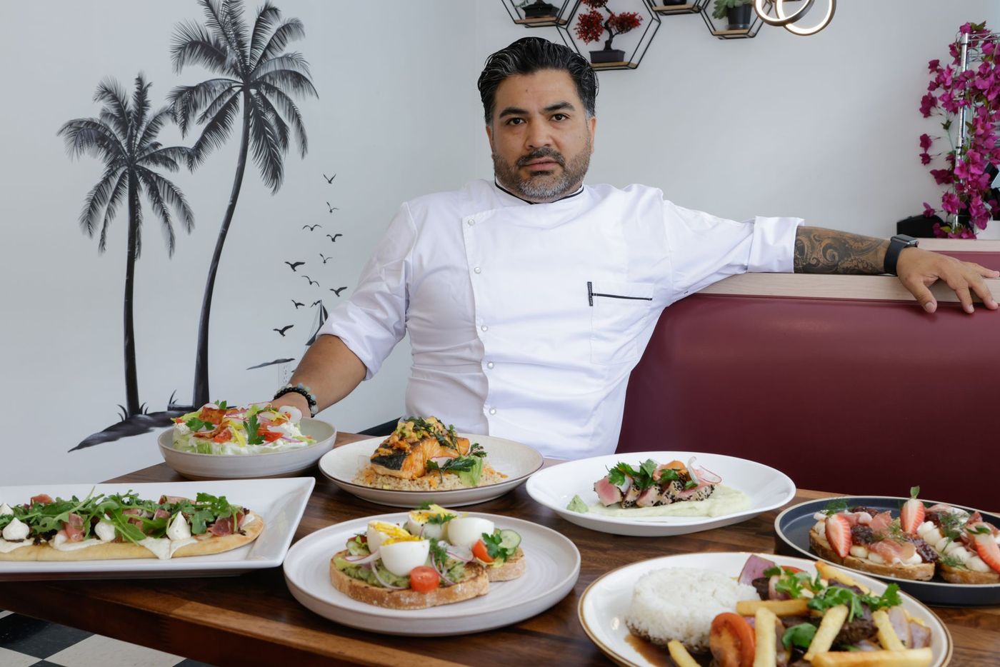 Chef in white coat, surrounded by dishes. Palm tree mural in background.