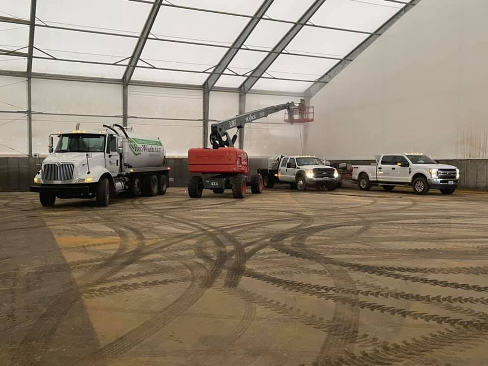 Trucks, tractor, and lift inside large tent; worker in lift, tire tracks on floor.