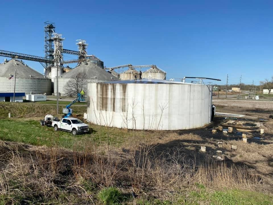 White truck and lift near a large, cylindrical concrete structure. Grain silos in the background. Blue sky.