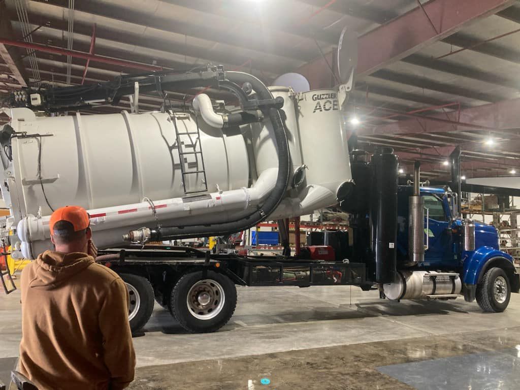 Man looking at a large, blue vacuum truck inside a warehouse. White tank, black hoses, red trim.