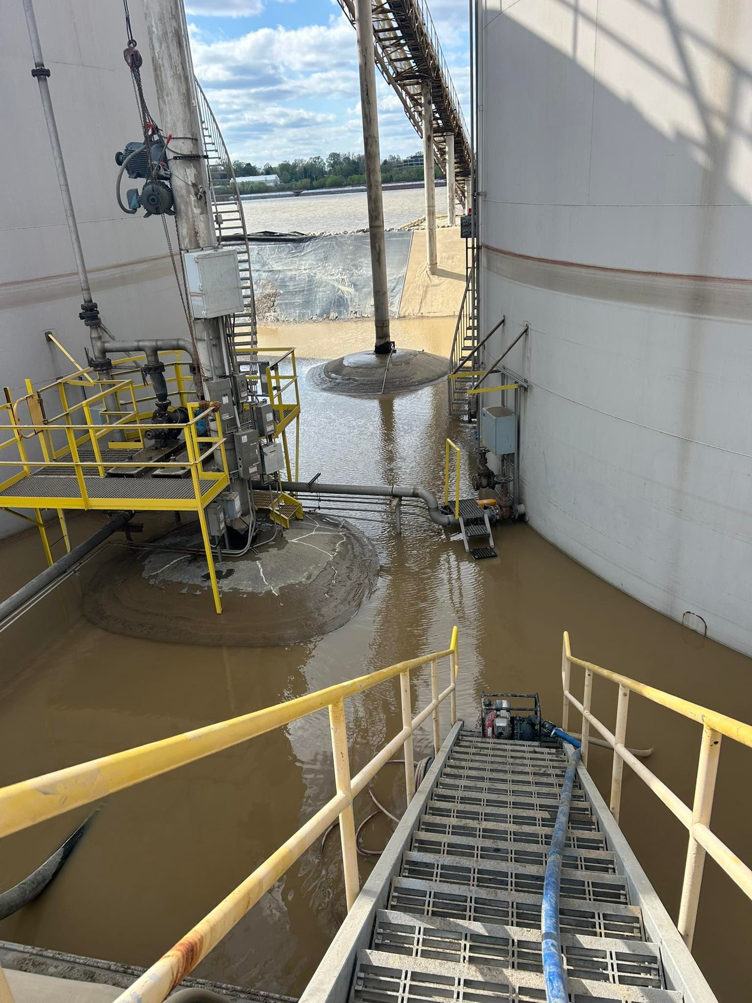 Flooded industrial area with stairs, yellow railings, and pipes submerged in brown water.