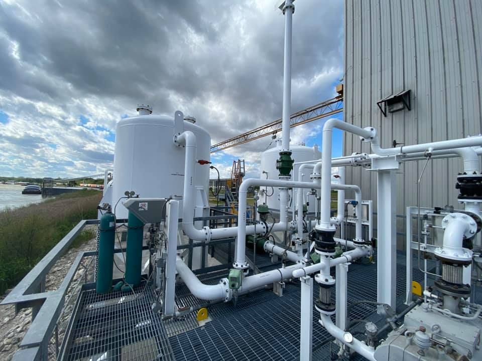 White industrial tanks and piping on a metal platform beside a building, under a cloudy sky.