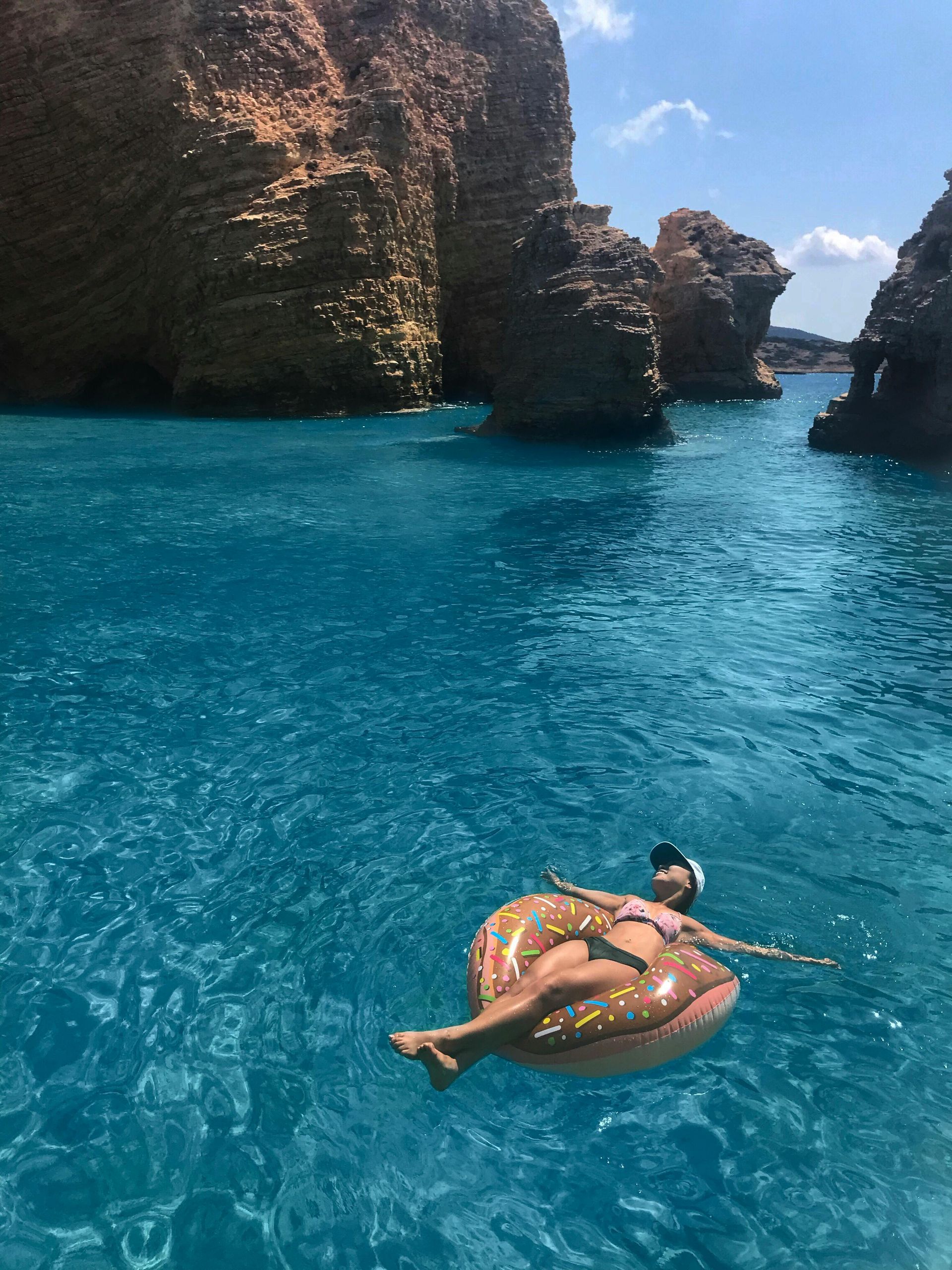 Woman floating on a donut-shaped inflatable in clear blue water surrounded by rock formations.