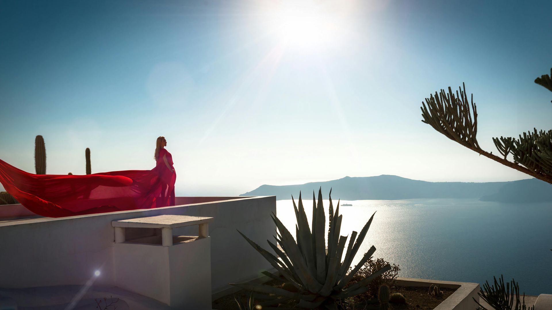 Woman in red dress on a terrace overlooking the Aegean Sea under bright sunlight.