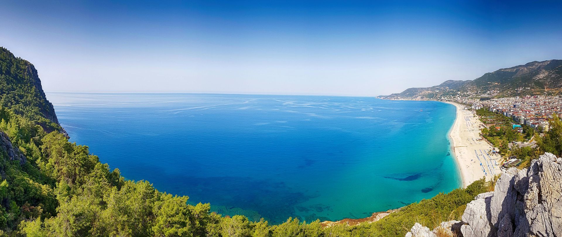 Vast turquoise water and a beach with a city, viewed from a high vantage point with lush greenery and rocky outcrops under a clear blue sky.