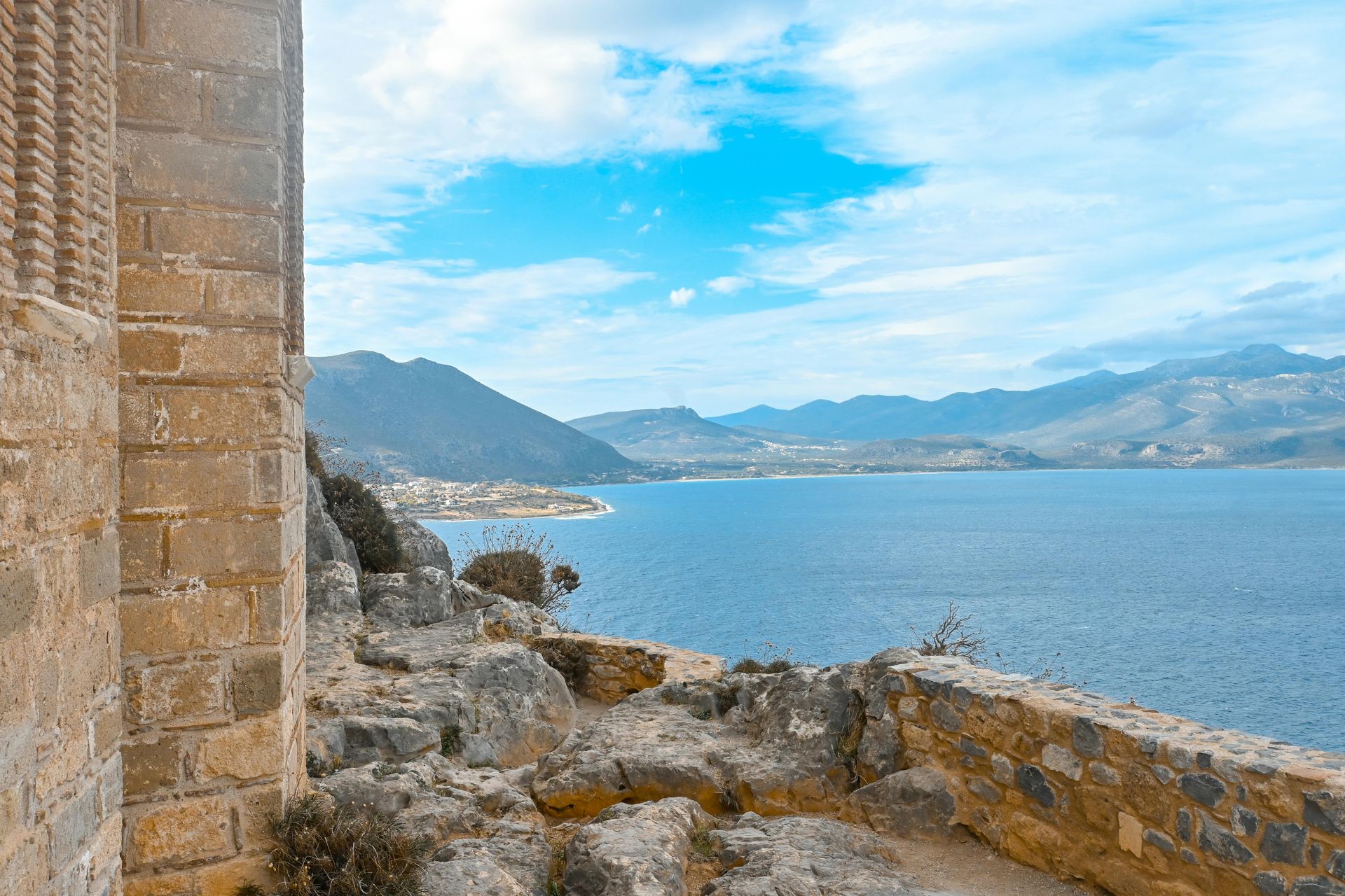 Stone structure overlooks blue sea, mountains, and sky.