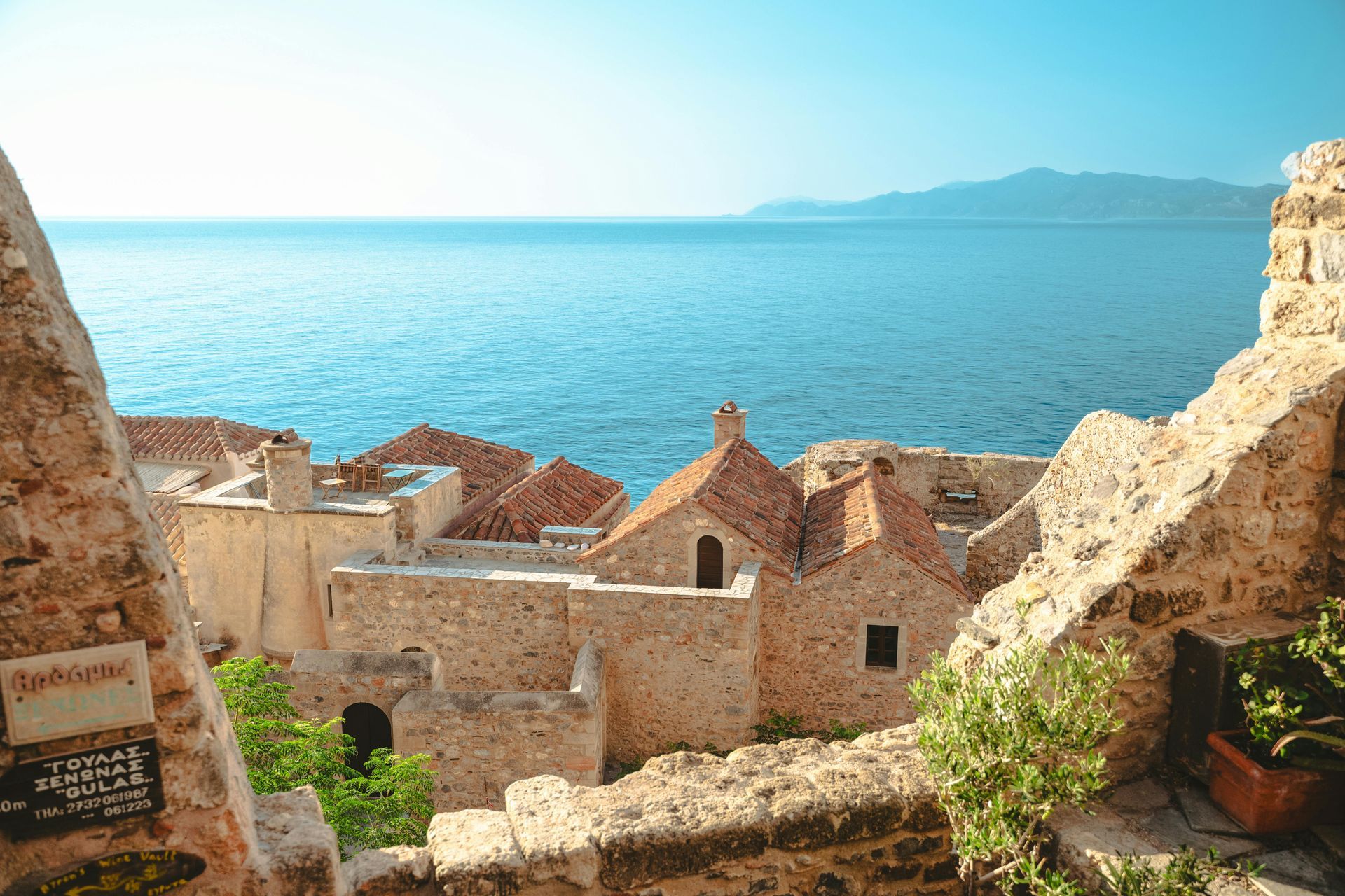 Stone buildings with red-tiled roofs overlooking blue sea, with a mountain in the distance.