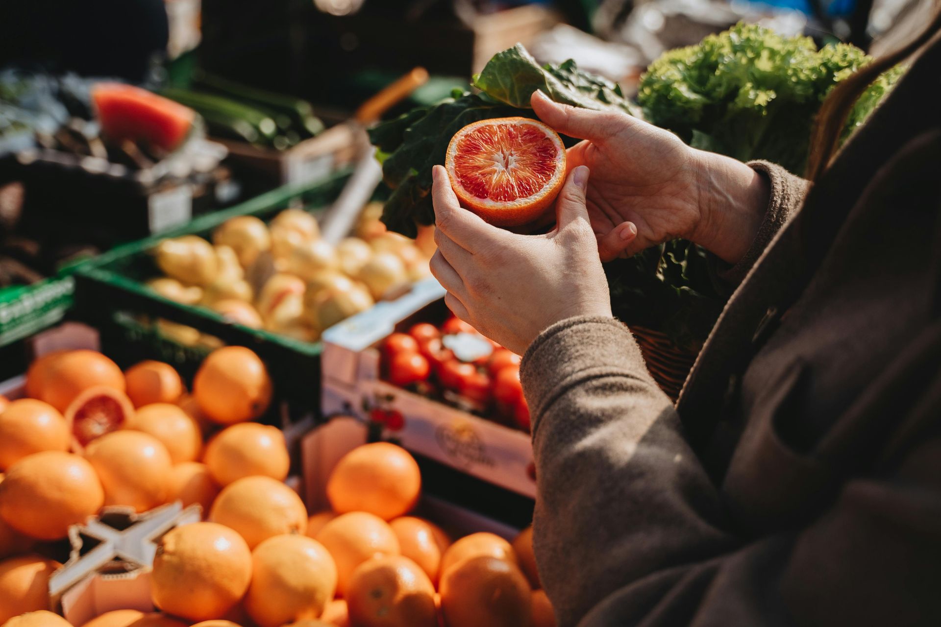 Person inspecting a halved blood orange at a produce market, surrounded by oranges and other fruits.