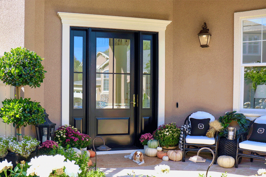 Black front door with sidelights, decorated porch with pumpkins, plants, and wicker chairs.
