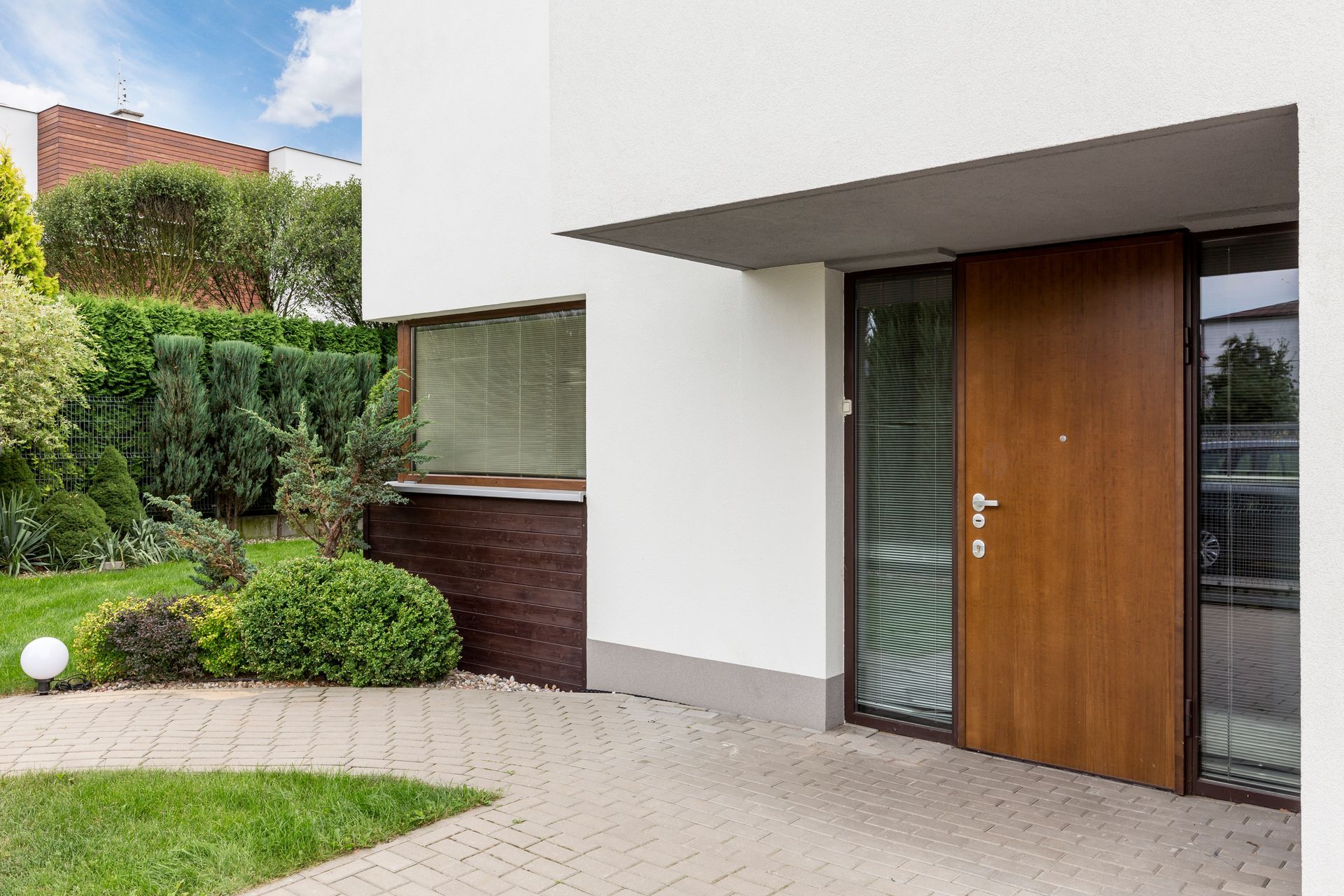 Modern house exterior with brown door and adjacent glass panel, paved walkway, and landscaping. Modern house exterior with brown door and adjacent glass panel, paved walkway, and landscaping.