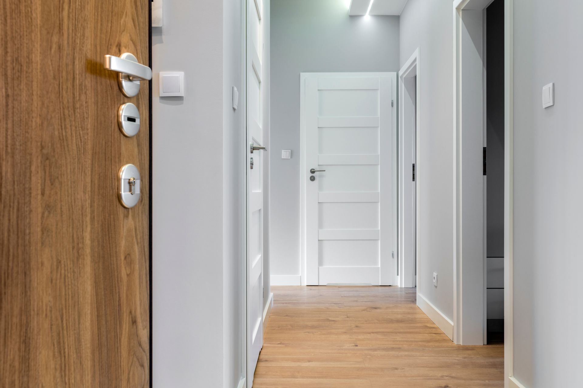 Hallway with wooden floor, light grey walls, and white doors. A wooden front door is on the left.