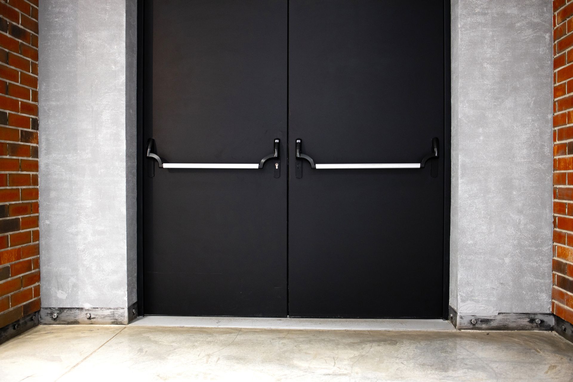 Black double doors with silver push bars, between brick and concrete walls. Black double doors with silver push bars, between brick and concrete walls.
