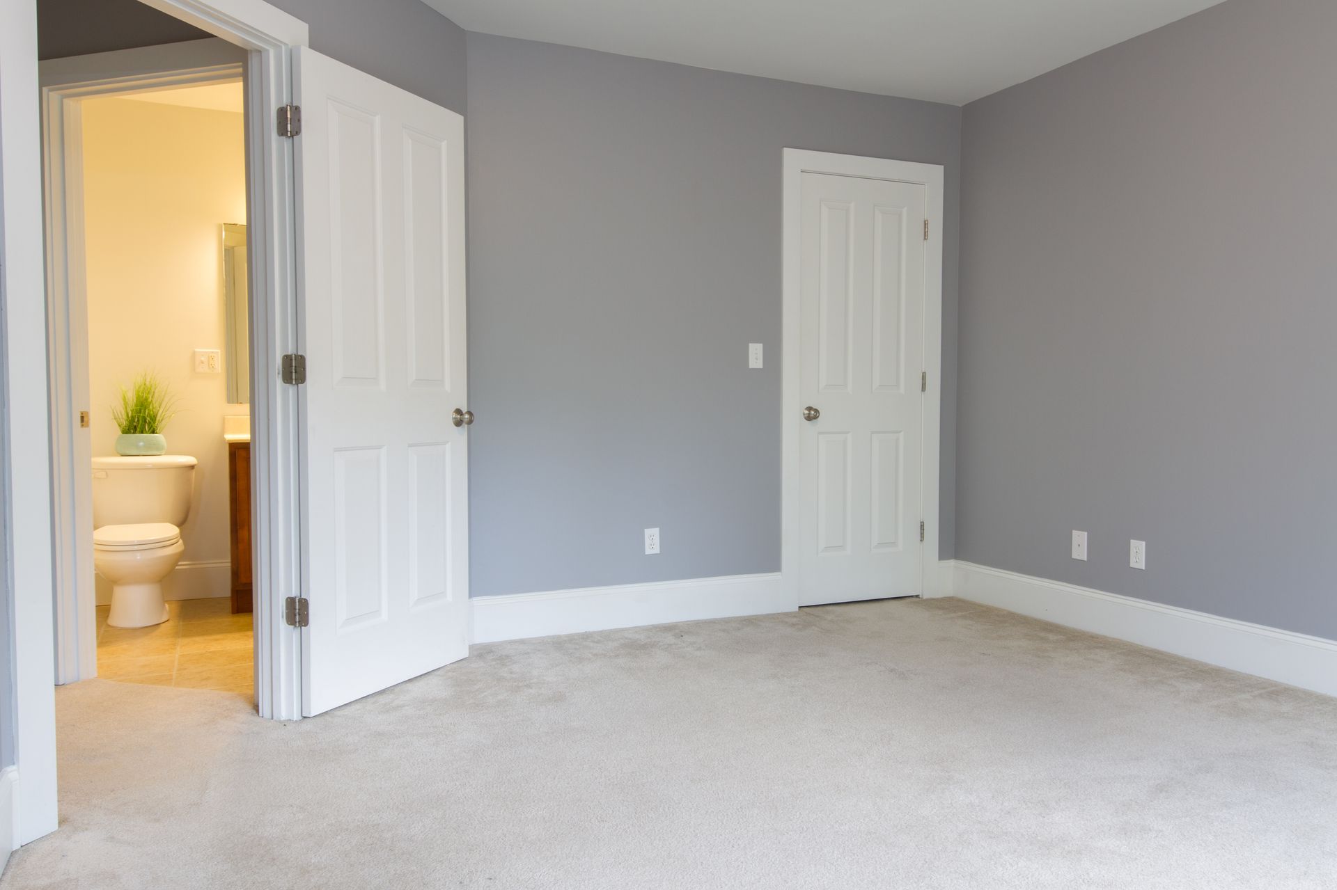 Empty room with grey walls, white doors, and a bathroom visible through an open doorway.