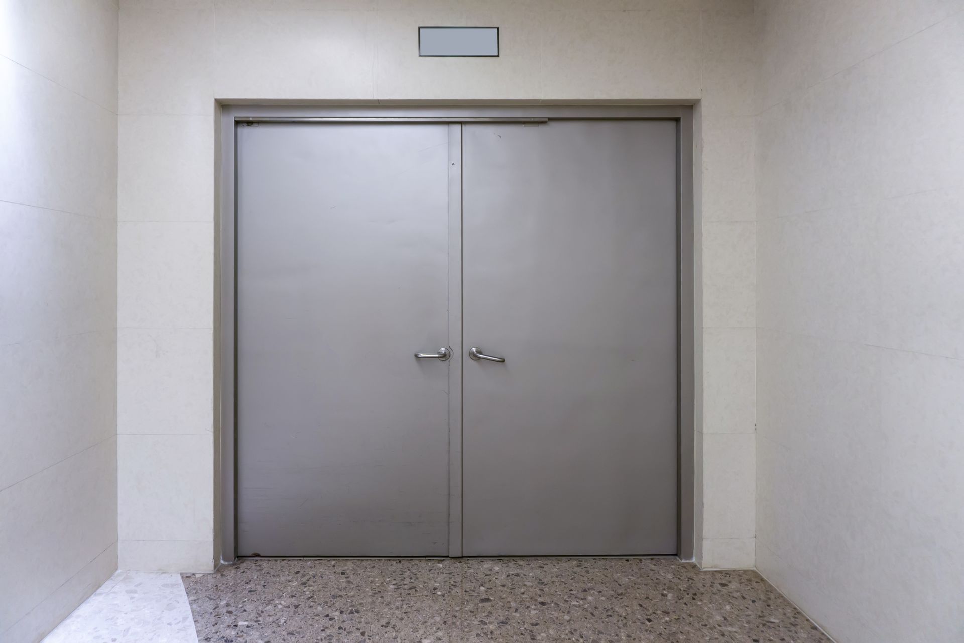Gray double doors in a white hallway with a textured floor and a rectangular light above. Gray double doors in a white hallway with a textured floor and a rectangular light above.