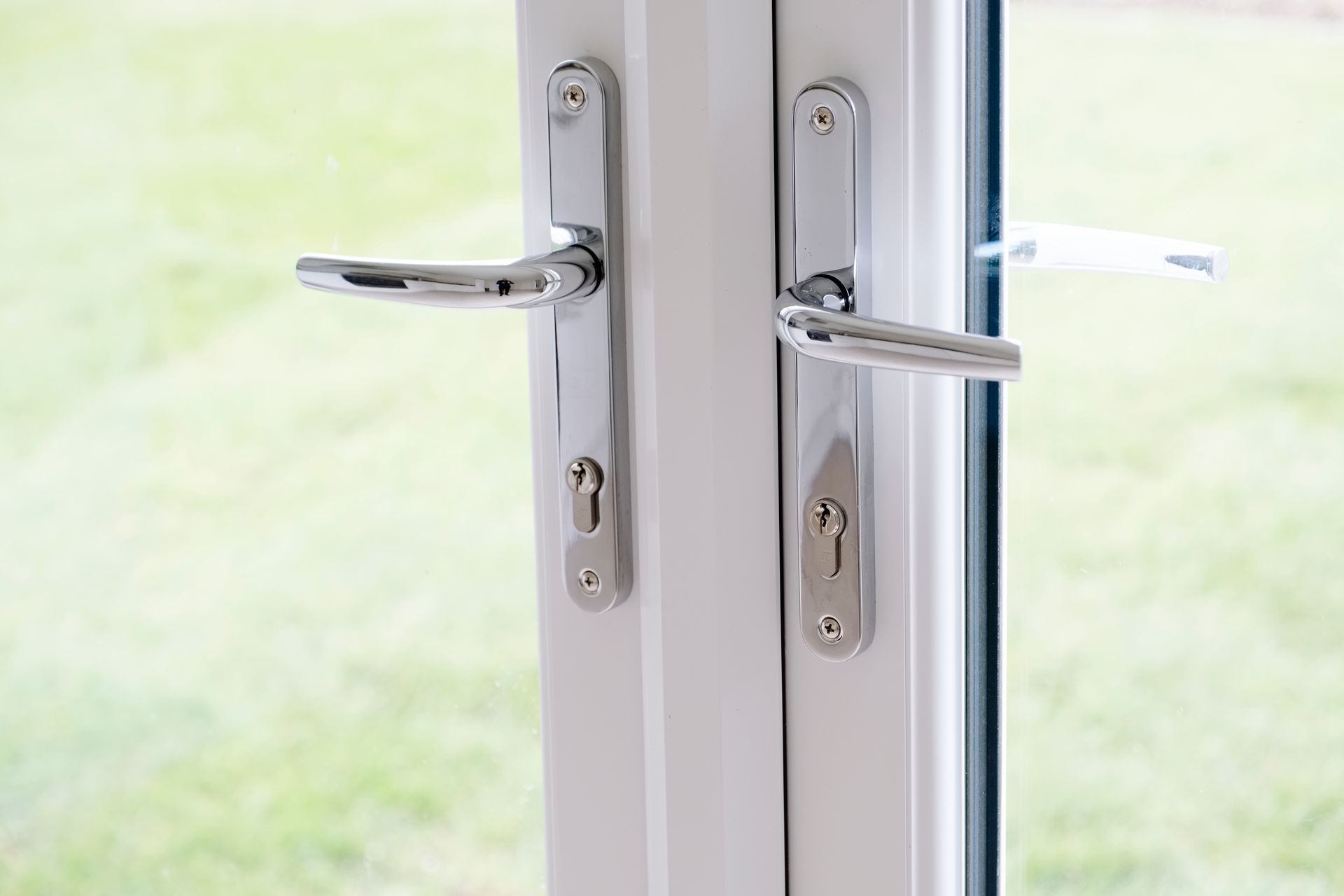 Close-up of white double doors with shiny silver handles and keyholes. Green lawn visible through the glass.