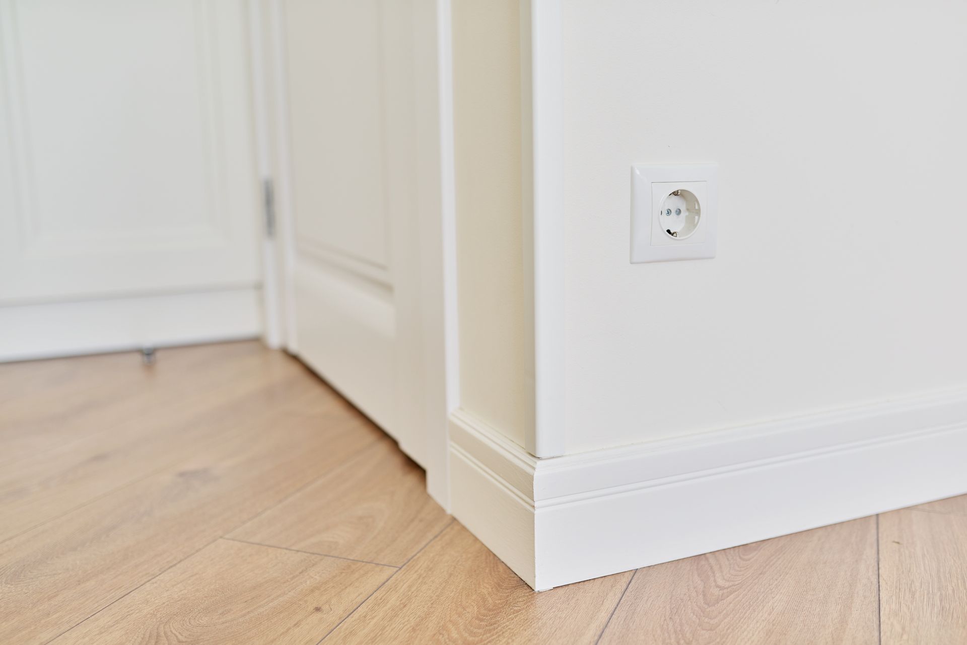 White wall corner with a power outlet, white door, and wooden floor.