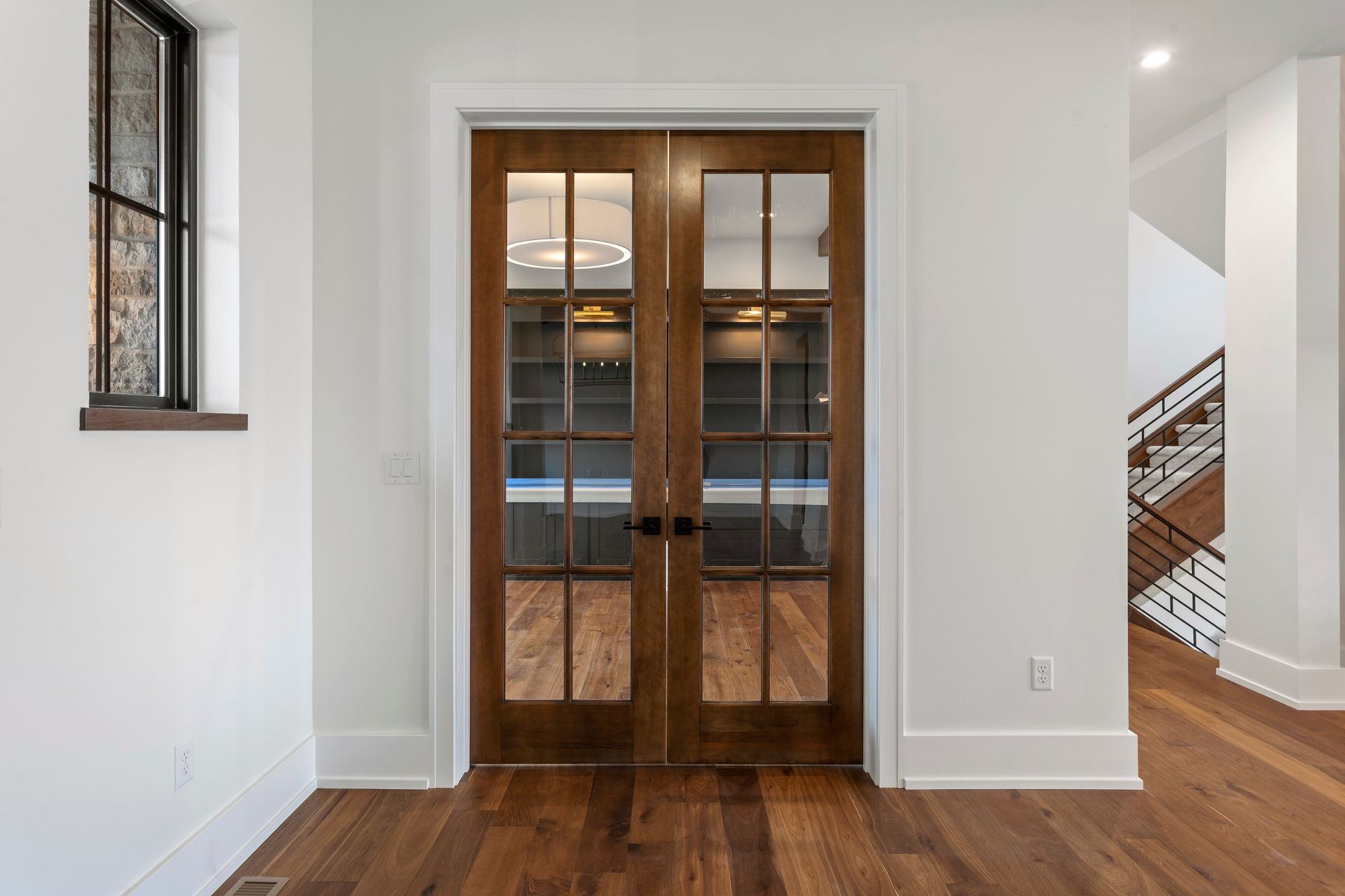 Double wooden doors with glass panes, set in a white-walled room, reflecting a room with shelving and a chandelier. Double wooden doors with glass panes, set in a white-walled room, reflecting a room with shelving and a chandelier.