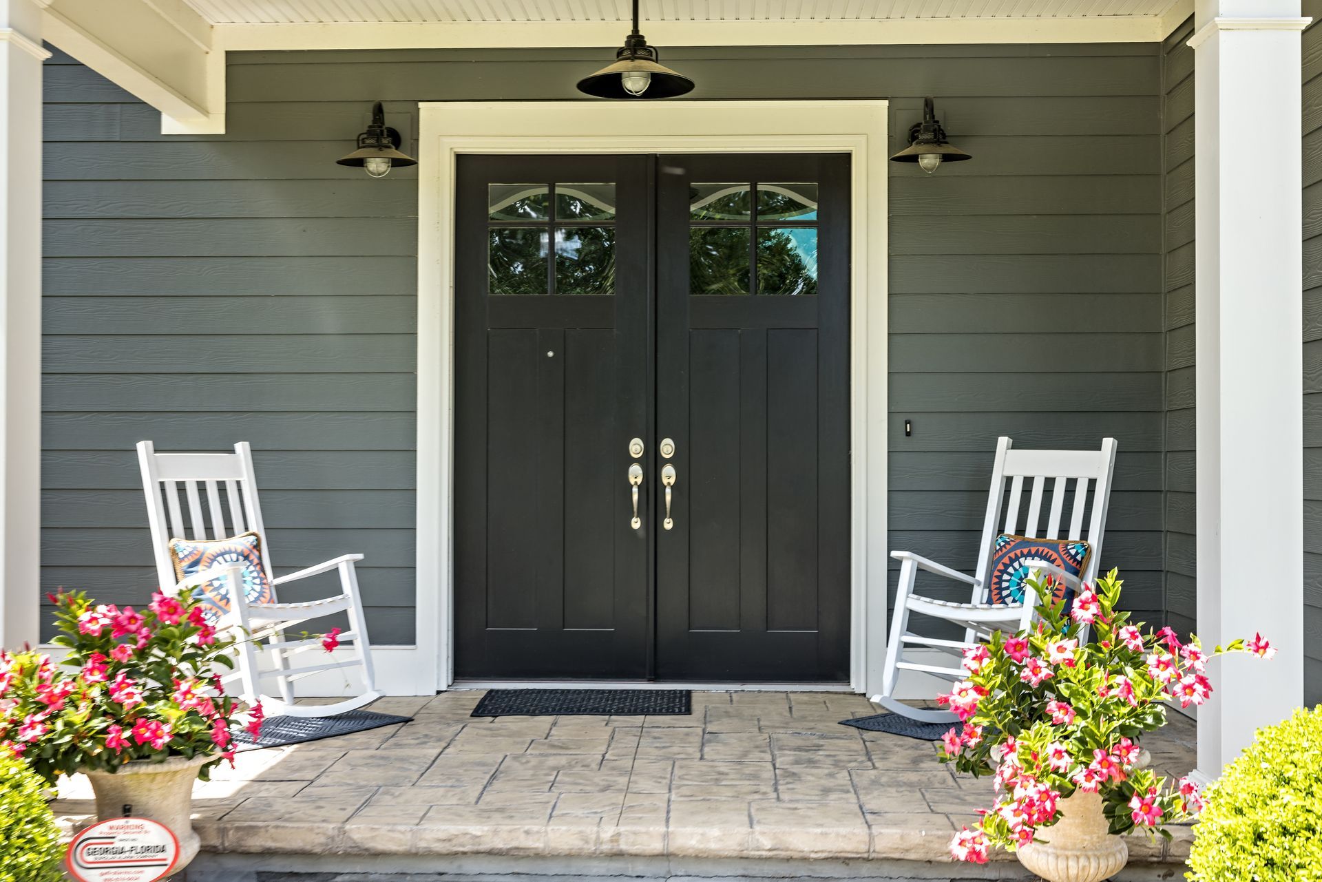 Gray house front with black double doors, white rocking chairs, and flower pots. Gray house front with black double doors, white rocking chairs, and flower pots.