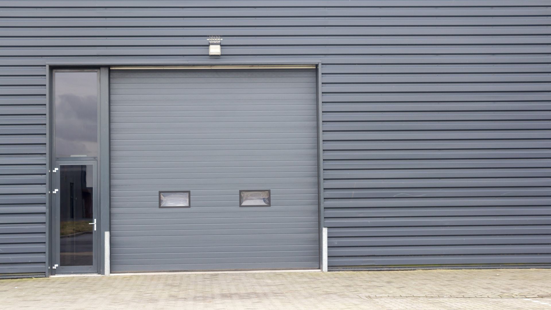 Gray industrial building with a closed garage door and a small open side door; a small light is above. Gray industrial building with a closed garage door and a small open side door; a small light is above.