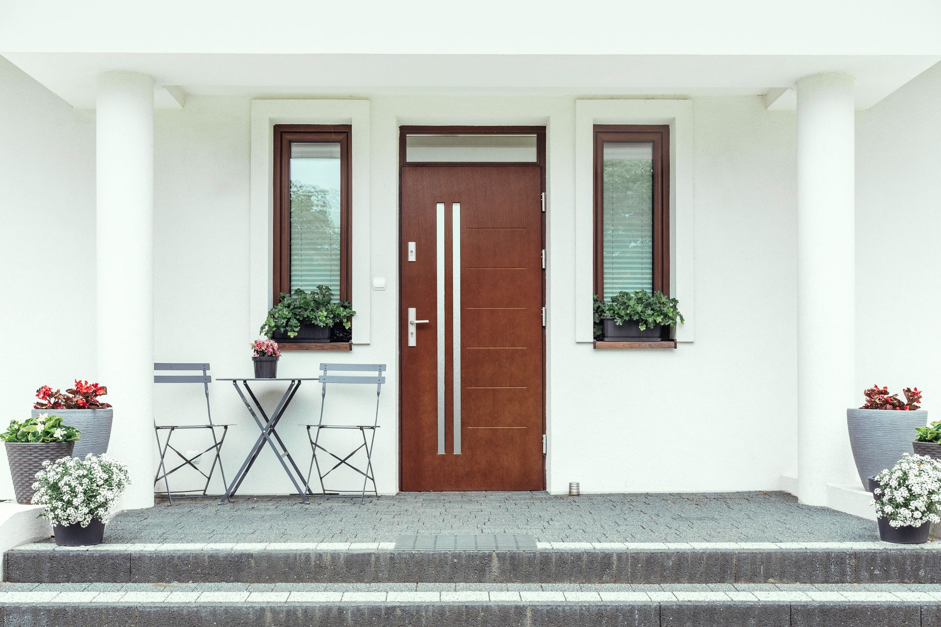 Brown door with silver handle centered between two windows and white pillars. Two chairs and a small table sit to the left. Brown door with silver handle centered between two windows and white pillars. Two chairs and a small table sit to the left.