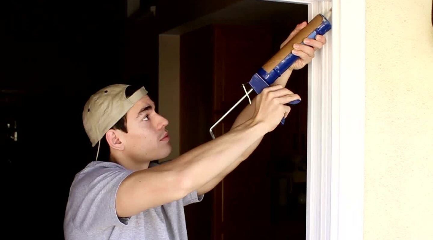 Man uses a caulk gun to seal a door frame, indoors.