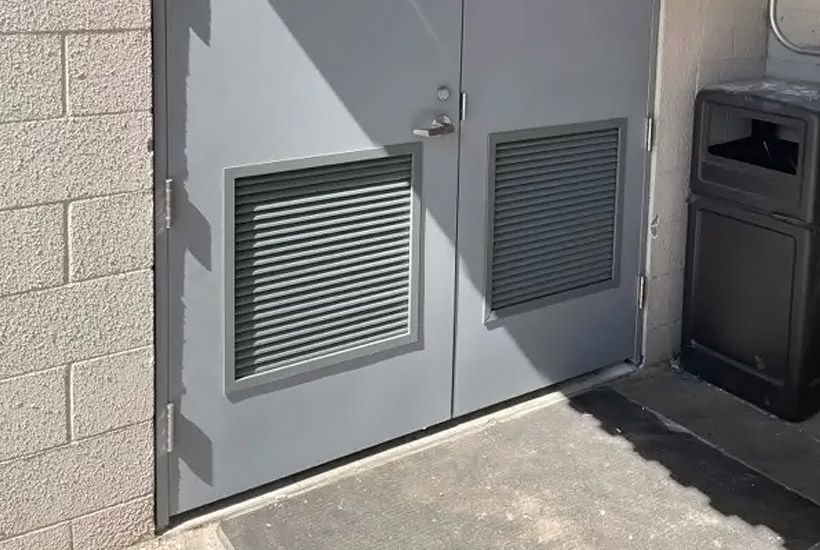 Gray double doors with ventilation grates, next to a brick wall and a trash receptacle. Gray double doors with ventilation grates, next to a brick wall and a trash receptacle.