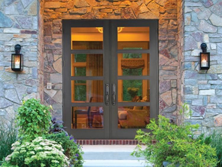 Double glass entry doors with dark frames set in a stone wall, flanked by lanterns and greenery.