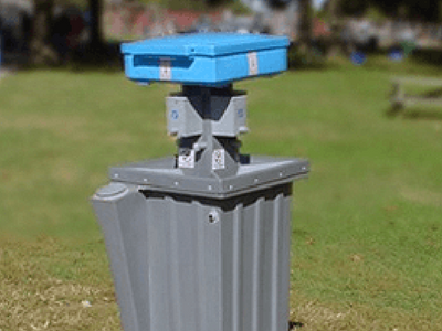 A gray outdoor waste bin with a blue, solar-powered compactor unit on top, situated in a green park.