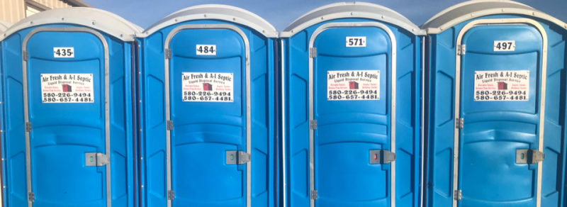 Four blue portable toilets lined up side-by-side with identification numbers and service stickers on the doors.