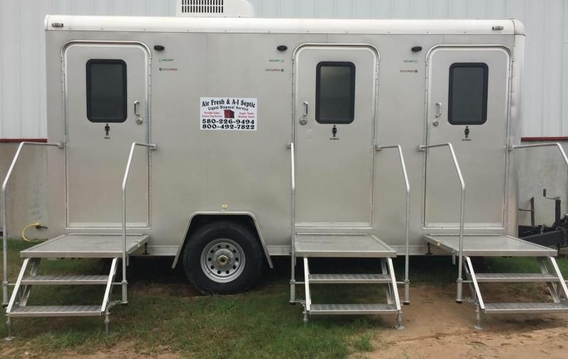 A portable three-stall restroom trailer with silver exterior, steps, and handrails, parked outdoors on grass.