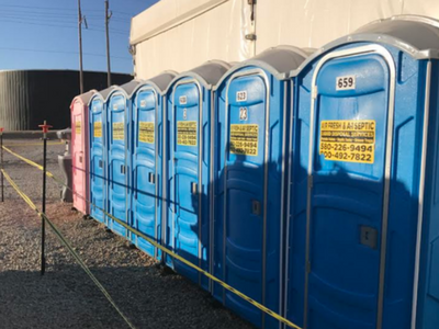 A row of blue portable toilets and one pink unit are lined up outdoors behind yellow caution tape.