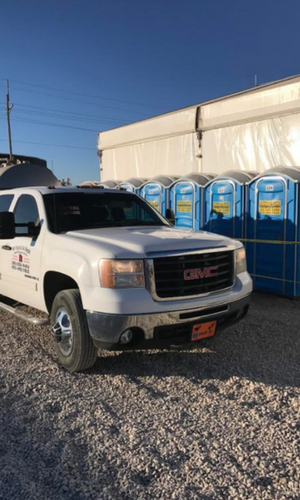 White GMC pickup truck parked on gravel next to a row of blue portable toilets.