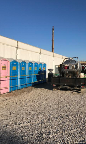 A row of blue portable toilets and one pink one stand on a gravel lot next to a sanitation truck under a clear blue sky.